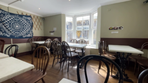 A room with pale walls, wooden floor, with wooden chairs, white tables, and a bay window on the far wall