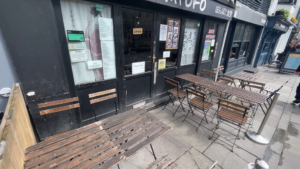 Outdoor seating consisting of wooden garden chairs and tables, on grey pavement, outside the black storefront