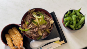 A bowl containing beef, rice, bell peppers, onion, with a bowl each side, one containing tempura fried prawns, and the other containing edimame beans. Chopsticks along the bottom of the tray that the bowls sit on.